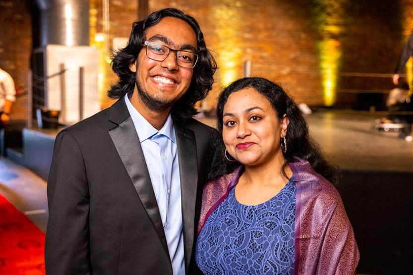 A man wearing a black blazer and light blue shirt smiles with a woman wearing a blue dress and purple wrap